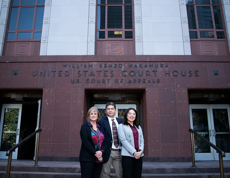 Three people standing in front of a brick building entrance with a sign above.